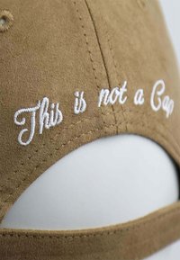 Braune Wildledermütze mit weißem gesticktem Text "Das ist keine Mütze." Nahaufnahme, die die Textur und die Nähdetails hervorhebt.