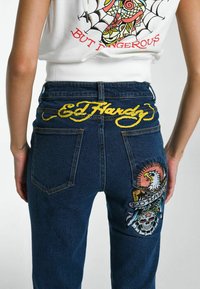 Jeans de mezclilla azul oscuro con el texto bordado en amarillo "Ed Hardy" en el bolsillo trasero, y coloridos diseños bordados de un águila y una calavera.