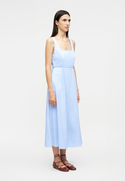 Femme aux longs cheveux foncés portant une robe midi sans manches à rayures bleues claires et blanches, et des chaussures plates lacées marron, posant devant un fond uni.