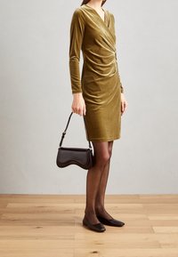 Robe en velours vert olive à manches longues avec un détail torsadé à l'avant. Associée à un sac à main noir et des ballerines marron. Collants noirs transparents.