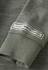 Hallivärvi sweatshirt'i kangas, kus lähedalt on nähtav varrukas, millel on musta värviga valgele taustale õmmeldud silt, millel on kirjas "KVALITEET".
