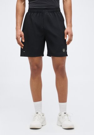 Bărbat purtând pantaloni scurți sport negri cu talie elastică, șosete albe până la gleznă și adidași albi, stând în picioare pe un fundal simplu.