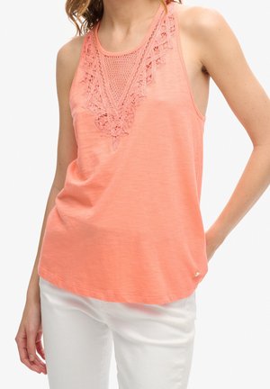 Kobieta ubrana w bezrękawnikowy koralowy top z koronkowymi i siateczkowymi detalami z przodu, w połączeniu z białymi spodniami, stoi z jedną ręką na biodrze.