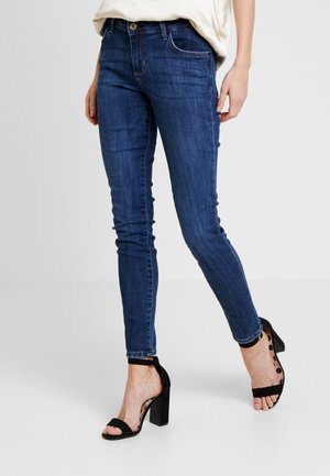 Personne portant un jean bleu slim et des sandales noires à talons hauts avec des brides à la cheville, debout devant un fond clair uni.