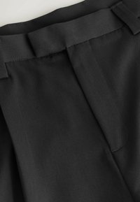 Gros plan sur un tissu noir montrant une ceinture avec passants et une poche latérale partiellement visible sur un pantalon ou une jupe.