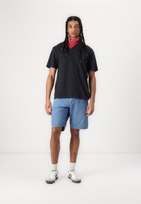 Zwarte korte mouwen V-hals t-shirt, denim shorts, rode bandana, witte sneakers met zwarte strepen en grijze zolen. Casual streetwear look.