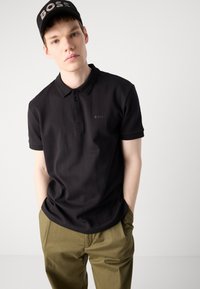 Čierna polo košeľa s jemnými vertikálnymi prúžkami, dvomi gombíkmi a logom "BOSS" na hrudi; v kombinácii s olivovo zelenými nohavicami.