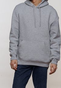 Sudadera gris con capucha ajustable, bolsillo canguro, puños y dobladillo de canalé. Tejido suave con una apariencia texturizada. Llevada sobre unos jeans oscuros.