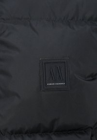 Black puffer jacket fabric with textured surface; features a rectangular black label with "AX" and "ARMANI EXCHANGE" in white text.