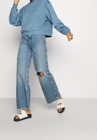 Sweat-shirt court bleu, jean large bleu clair déchiré avec ourlet effiloché, et sandales blanches avec semelles noires et boucles argentées.