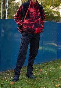 Fleece mikina s červeno-černým vzorem a půlzipem, spárovaná s černými cargo kalhotami a černými botami, na modrém pozadí a trávě.