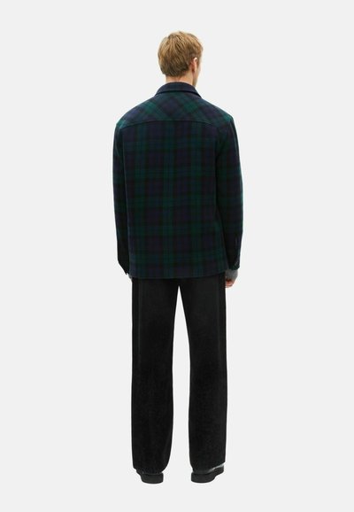Férfi áll, hátat fordítva, sötétzöld és tengerészkék kockás zakót, fekete, bő szárú nadrágot és fekete cipőt viselve, fehér háttér előtt.