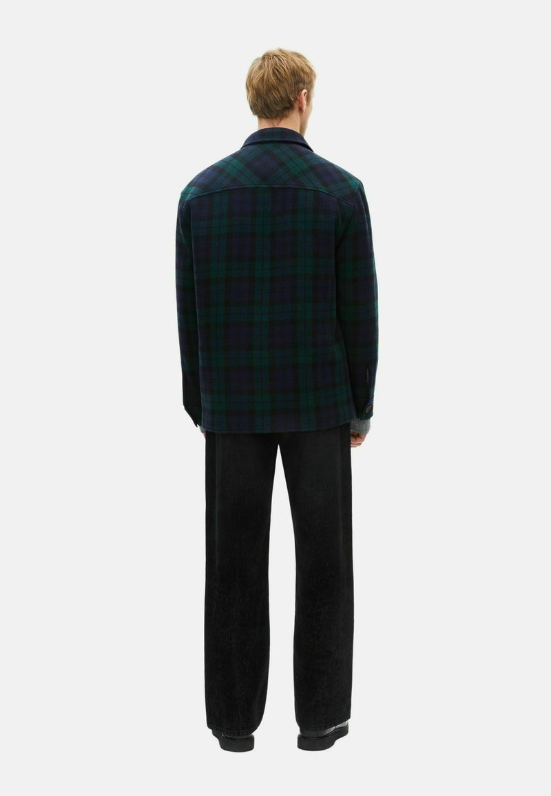 Mann steht mit dem Rücken zur Kamera, trägt eine dunkelgrüne und marineblaue karierte Jacke, schwarze weite Hosen und schwarze Schuhe vor einem weißen Hintergrund.