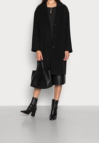 Czarny wełniany płaszcz z wyłogowym kołnierzem i guzikami z przodu, zestawiony z czarną skórzaną torbą typu bucket i dopasowanymi skórzanymi botkami za kostkę.