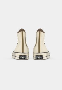 Zapatillas de altura alta en lona beige con acentos marrones, suela de goma blanca y parche de logo negro en el talón. Strips verticales en la parte posterior.