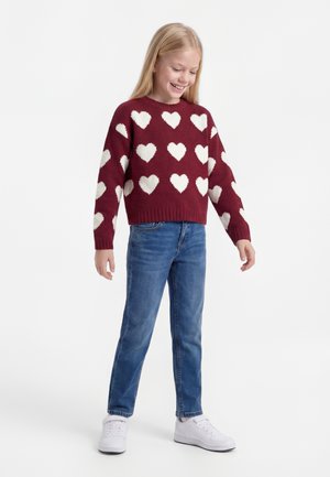 Smilende jente står iført rød genser med hvite hjerter, blå jeans og hvite joggesko mot en ensfarget lys bakgrunn.