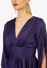 Abito viola profondo con scollatura a V, maniche lunghe e fluide, e vita arricciata. Tessuto satinato liscio con una leggera lucentezza.