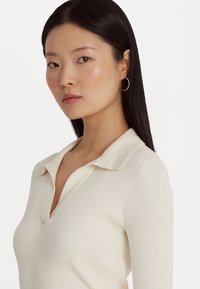 Ριχτή, κρέμα polo πουλόβερ με V-λαιμόκοψη και μαλακή λεπτομέρεια στον γιακά. Το μοντέλο φορά χρυσά σκουλαρίκια κρίκους. Απλό λευκό φόντο.