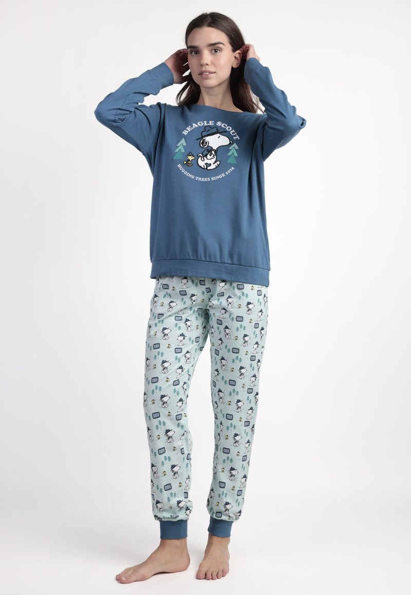 Joven mujer ajustándose el cabello, vestida con un sudadera azul de "Beagle Scout" y pantalones de pijama de patrón de Snoopy en azul claro, de pie descalza sobre un fondo blanco.