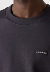 Granatowy sweatshirt z prążkowanym kołnierzem, o miękkiej fakturze, z czarną prostokątną naszywką z logo "Calvin Klein" na lewej stronie klatki piersiowej.