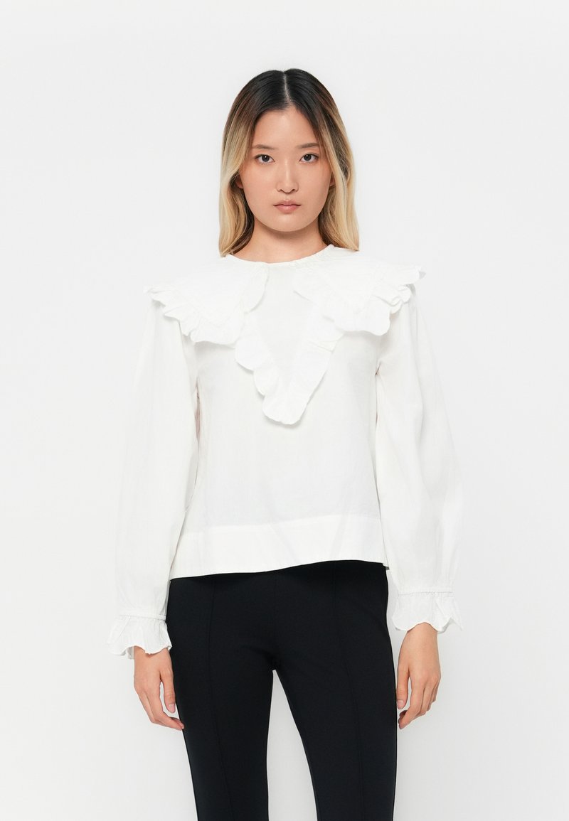 Blusa bianca con colletto e polsini a volant, maniche lunghe e vestibilità ampia. Presenta una texture di tessuto e un design semplice e minimalista.