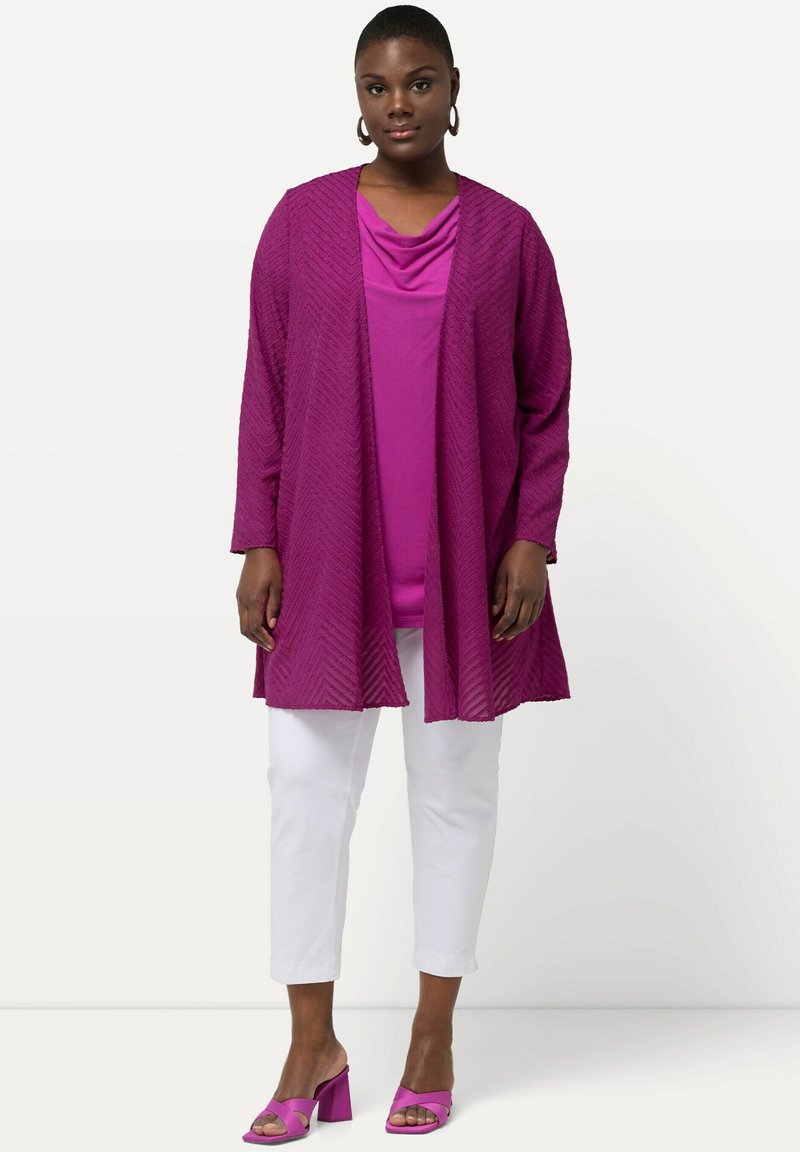 Magenta kardigan ve stylu kimona s texturovaným vzorem chevron, vrstvený přes odpovídající top s rolákem, bílé krátké kalhoty a růžové sandály na blokovém podpatku.