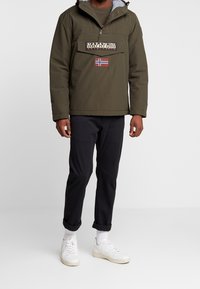 Oliwkowa bluza z kieszenią z przodu, z naszywką logo i norweską flagą. W połączeniu z podwiniętymi czarnymi spodniami i białymi sneakersami.