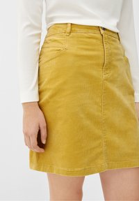 Gul kordskjol med fickor, med en figurnära midja och rak skärning, visas med en vit långärmad topp.