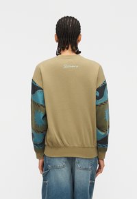 Άτομο με πλεξούδες που φοράει φούτερ ελιάς με μανίκια σε μπλε και μαύρο αφηρημένο μοτίβο και ανοιχτόχρωμο τζιν παντελόνι.