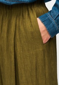 Olivgrüne, plissierte Stoffhose mit strukturierter Oberfläche, seitlichen Taschen und einem am Rand sichtbaren denimblauen Hemdblusenbund.