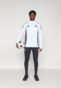 Športnik, oblečen v belo majico z dolgimi rokavi s črnimi in vijoličnimi poudarki, črne oprijete hlače, drži nogometno žogo, stoji na nevtralnem ozadju.