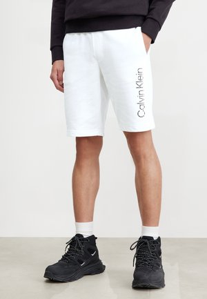 Mężczyzna noszący białe szorty Calvin Klein z pionowym logo na lewej nodze, czarną bluzę, białe skarpetki i czarne buty trekkingowe, stojący.