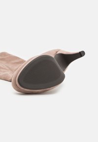 Różowe zamszowe buty na obcasie z trójkątną czarną gumową podeszwą i teksturowanym gripem, charakteryzujące się eleganckim, spiczastym czubkiem.