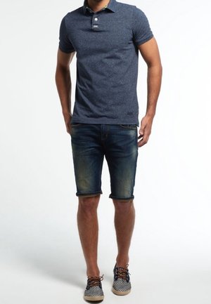 Homme portant un polo bleu marine, un short en denim délavé et des baskets à lacets à motifs, se tenant les mains près des hanches.