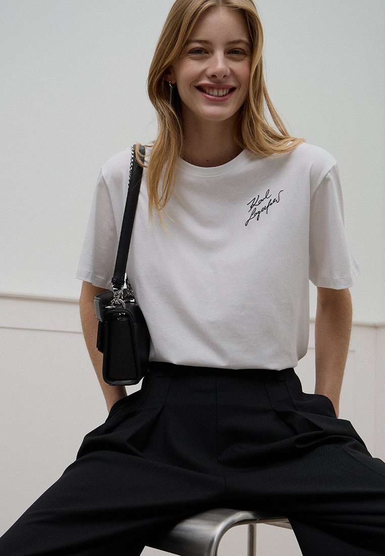 Weißes Baumwoll-T-Shirt mit kurzen Ärmeln, mit einem schwarzen handgeschriebenen Logo auf der Brust, kombiniert mit hochgeschnittenen schwarzen Hosen.