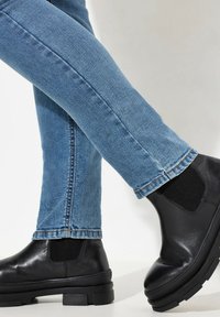 Botas Chelsea negras de cuero suave con paneles laterales elásticos, combinadas con jeans ajustados azules, mostradas desde el tobillo hacia abajo.