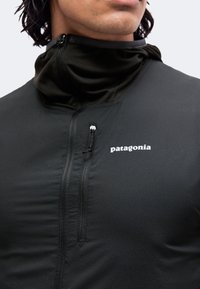 Άνδρας που φοράει μαύρο μπουφάν Patagonia με φερμουάρ και ανασηκωμένο γιακά, εμφανίζοντας μερικό πρόσωπο και λαιμό σε απλό ανοιχτό φόντο.