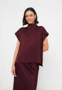 Haut oversize bordeaux avec col montant, manches courtes et coupe décontractée, réalisé en tissu lisse, associé à une jupe assortie.
