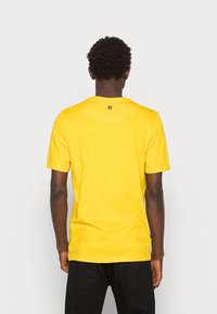 Osoba o ciemnej karnacji ubrana w jasną żółtą koszulkę z krótkim rękawem i czarne spodnie, pokazana od tyłu na jednolitym tle.