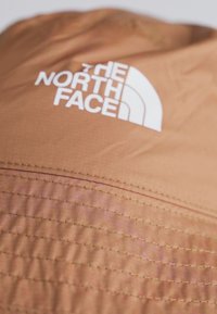 Brązowy materiałowy kapelusz z przeszywaną teksturą, z wyraźnym białym logo "The North Face" z przodu.
