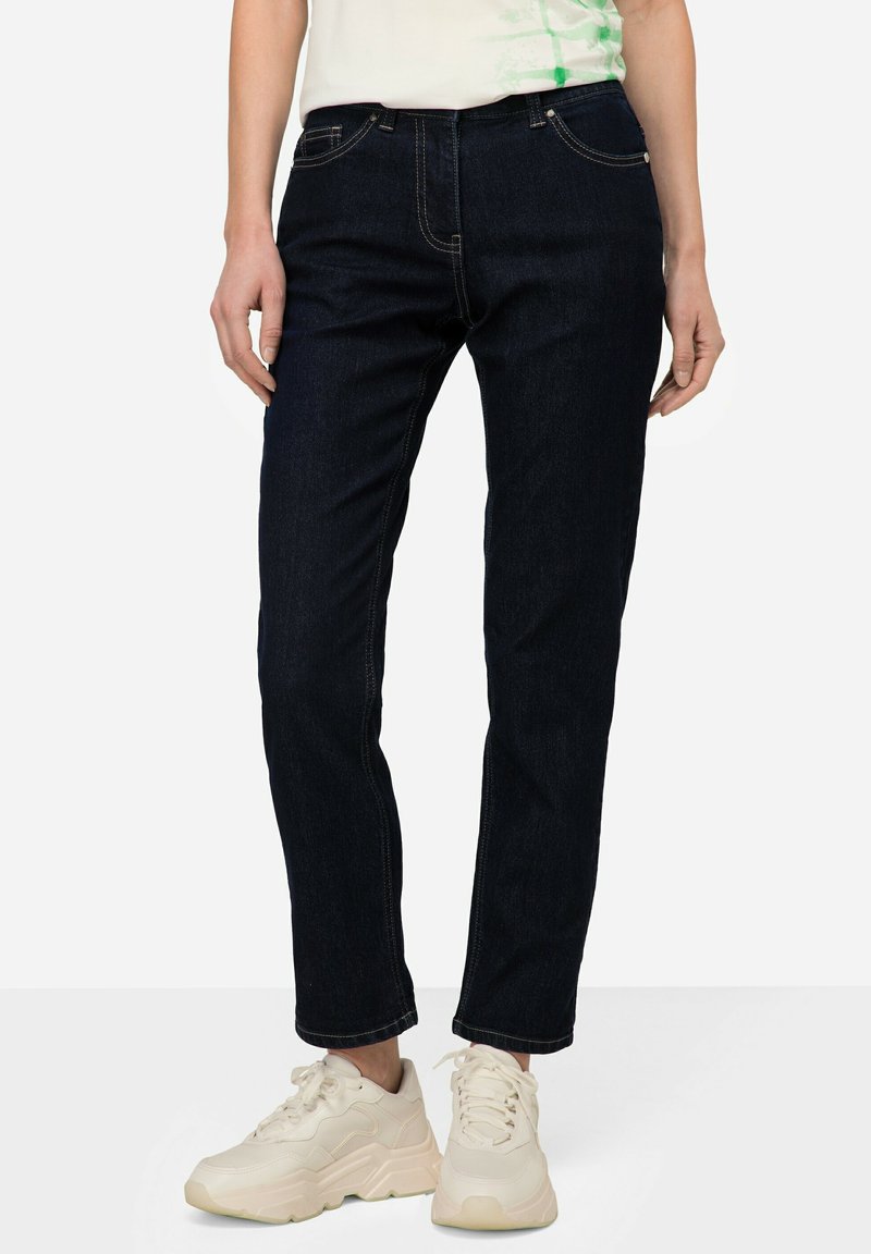 Jeans de mezclilla azul oscuro con corte slim, diseño de cinco bolsillos y costuras en contraste. Llevados con zapatillas gruesas de color claro.