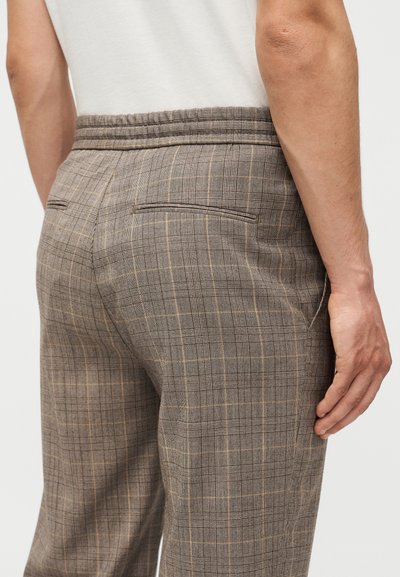 Η όψη από πίσω ενός ατόμου που φοράει μπεζ και καφέ καρό παντελόνια με πίσω τσέπες με καπάκι και ελαστική μέση, σε συνδυασμό με ένα λευκό πουκάμισο.