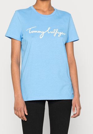 Personne portant un t-shirt bleu clair à manches courtes avec un texte blanc "Tommy Hilfiger" et un pantalon noir, montrée du cou aux hanches.