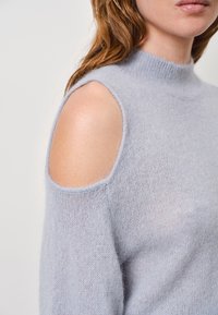 Lichtblauwe gebreide trui met een hoge kraag, met uitsneden op de schouders en een zachte, fuzzy textuur. Naaddetails zichtbaar op de mouwen.