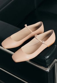 Pereche de balerini roz nude cu vârfuri pătrate și curele cu cataramă așezați pe brațul unei canapele din piele neagră.