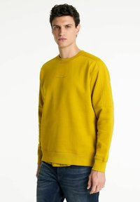 Sudadera amarilla con cuello redondo, puños y dobladillo acanalados. Presenta un sutil detalle de logo en el pecho. Llevada sobre jeans azules.