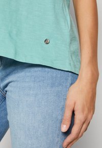 T-shirt en coton vert menthe avec une légère texture, un ourlet arrondi, des manches courtes et un accent logo argenté en bas à gauche. Associé à un jean bleu clair.