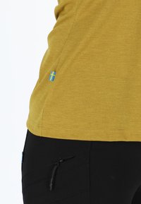 Sennepsgul skjorte med struktureret stof, der har en lille blå-og-gul svensk flag accent på venstre side nær kanten.