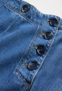 Denimplagg har ett blått tyg med synlig textur, sex svarta knappar, gul söm och en lätt samling upptill.