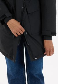Zwarte buitenjacket heeft een ritssluiting, drukknoopsluiting en twee voorzakken. Het materiaal lijkt glad met een lichte glans.